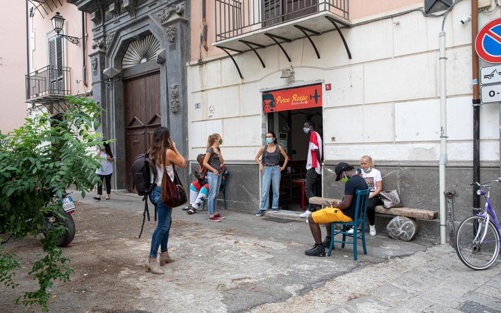 5 Personen stehen vor dem Eingang zum Zentrum von Arci Porco Rosso. Zwei weitere Personen sitzen, eine auf einer Bank und eine auf einem Stuhl. Am linken Rand des Bildes befindet sich eine größere Pflanze.