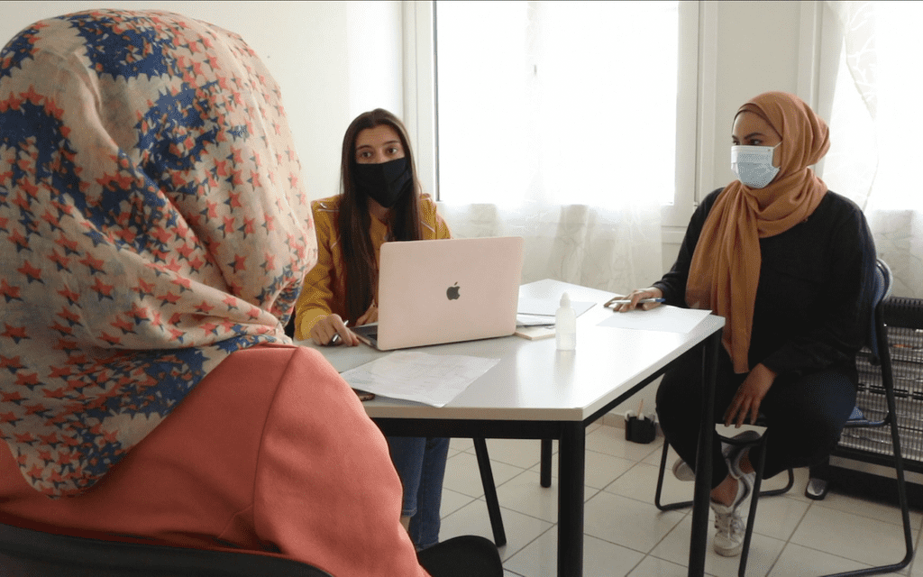 Three women sit around a table. Two women wear headscarfs. On the table is a laptop from Apple.