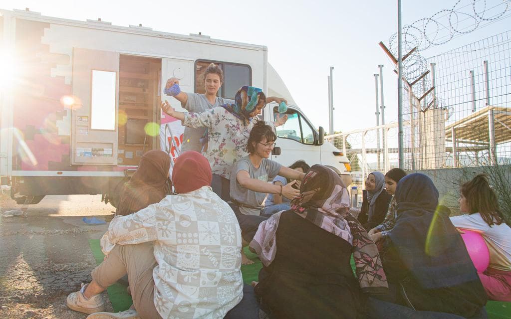 A handful of women, some with children, are sitting in front of a white van. Two women are standing seemingly explaining something. In the background, on the left side of the picture, the sun shines.